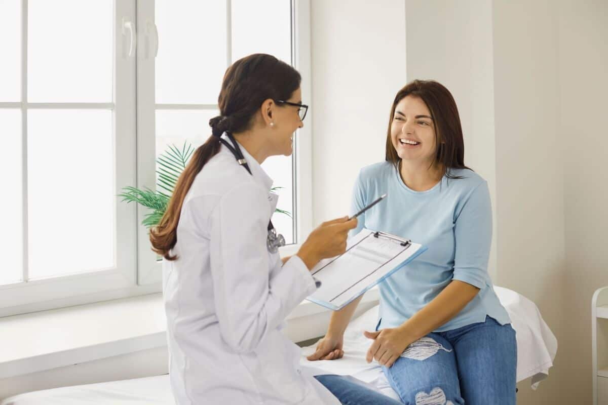 médica e paciente sentadas em maca. a médica veste jaleco branco e segura prancheta e caneta. a paciente sorri e veste blusa azul-claro